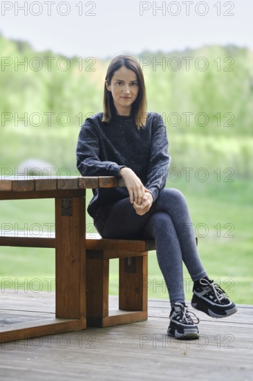 A young woman sits on a wooden bench beside a table, wearing casual attire. She enjoys the peaceful outdoor environment surrounded by greenery. Soft daylight enhances the serene atmosphere
