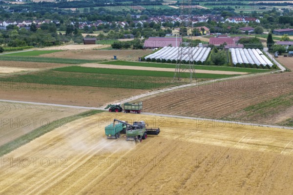 A combine harvester and a tractor work on a harvest field near a village with greenhouses