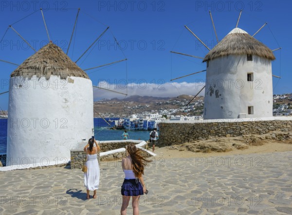 Mykonos, Cyclades, Greece - Tourists photograph the six sixteenth-century windmills lined up on a hill above Mykonos Town, Mykonos Chora. The windmills are the island's landmark. At the back, right by the sea, are the colorful houses of Little Venice. Mykonos is part of the Cyclades archipelago in the Aegean Sea