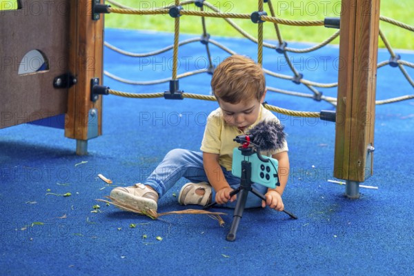 Little child sitting on playground's rubberized floor setting up a smartphone on a tripod with a microphone attached, creating videos and digital content
