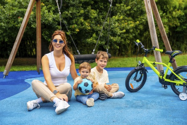 Happy mother sitting with her two sons on a playground's blue rubberized surface near a bicycle and a swing, enjoying a sunny day outdoors after playing with a ball