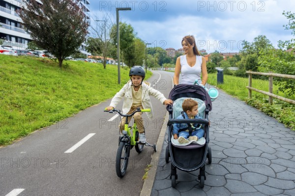 Mother enjoying quality time with her two sons in a serene urban park, one riding a bicycle with training wheels and the other in a stroller, promotes family bonding