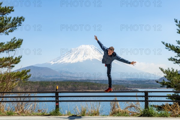 Young man balancing on railings next to a road, view across the lake to Mt Fuji volcano, Motosu Lake, Yamanashi Prefecture, Japan