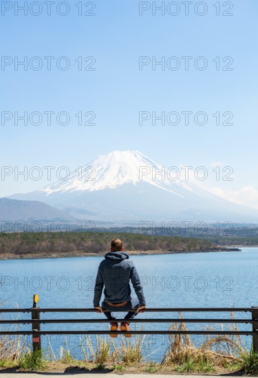 Young man sitting on railings next to a road and looking across the lake to Mt Fuji volcano, Motosu Lake, Yamanashi Prefecture, Japan