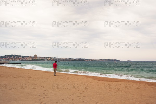 Man wearing a red shirt looking at the moving sea, Fréjus in the background, Provence-Alpes-Côte d'Azur region, France