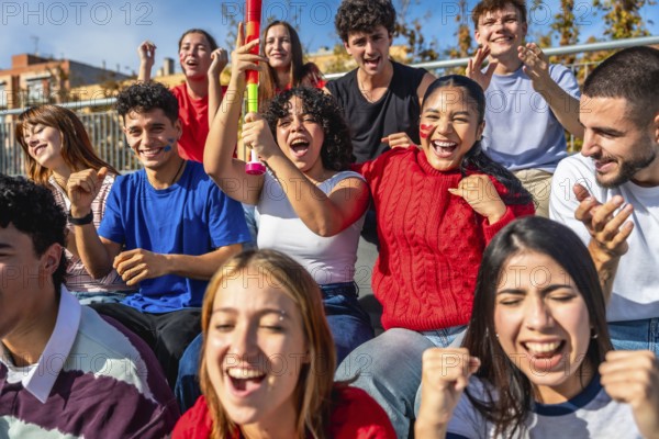 Diverse young friends on stadium bleachers, cheering, clapping and raising hands in daylight as they passionately support their team, smiling and celebrating together during a game