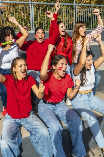 Young women sitting on stadium bleachers, cheering loudly with raised arms and painted faces, expressing excitement and support for their favorite team during a match