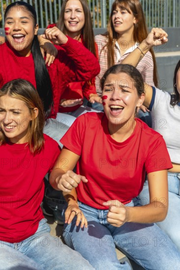 Group of enthusiastic young women friends wearing red shirts and face paint, actively cheering and expressing excitement while sitting together at a sports stadium