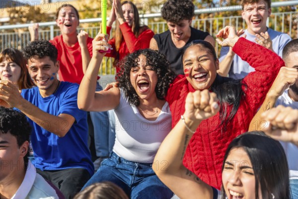 Diverse group of enthusiastic young people celebrating their team's victory at a stadium, waving hands, cheering loudly, and displaying team spirit with painted faces