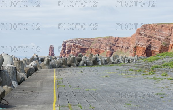 Coastline with concrete tetrapods by the sea, coastal protection, protective wall, paved coastal strip with yellow line, danger, marking, warning, red cliffs and Lange Anna in the background, cliff, cliffs, rock layers in red sandstone, surf pillar, escape line, perspective, overcast sky, blue sea on the horizon, west cliff, high-seas island of Heligoland, North See, Pinneberg District, Schleswig-Holstein, Germany
