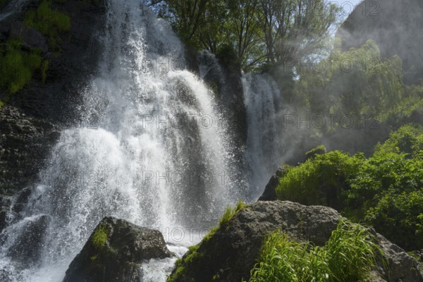 An impressive waterfall falls over rocks surrounded by green vegetation and trees in sunlight, Shaki Waterfall, Syunik Province, Syunik, Armenia