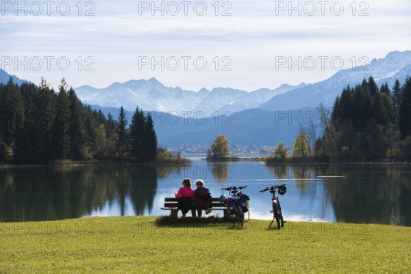 Two woman sitting on a bench look at the peaceful Forggensee with mountain views, surrounded by trees and bicycles, near Roßhaupten, Ostallgäu, Allgäu, Bavaria, Germany