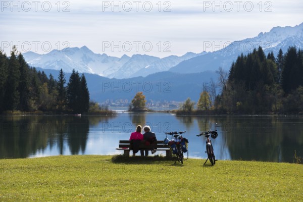 Two people sitting on a bench enjoying the view of the peaceful Forggensee and the majestic mountains in the background, near Roßhaupten, Ostallgäu, Allgäu, Bavaria, Germany