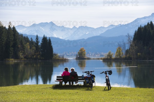 People sitting on a bench with a view over Lake Forggensee to Bergen, surrounded by peaceful nature and bicycles, near Roßhaupten, Ostallgäu, Allgäu, Bavaria, Germany
