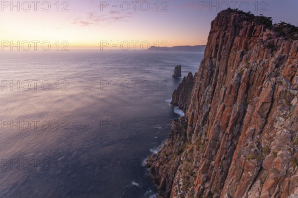 Long exposure at sunrise shows glowing dawn over the dramatic rocks on the Cape Hauy Track. Gentle waves hit the steep cliffs. Cape Hauy, Tasman Peninsula, Tasmania, Australia