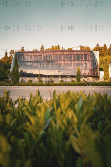 Glass façade of a modern building surrounded by trees in warm sunlight, green leaves foreground, Altensteig, Calw district, Germany