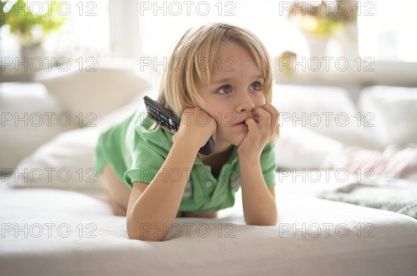 Child, boy, 7 years old, blonde, lying on sofa with remote control in hand, watching TV, watching television in front of television, excited, fascinated, Stuttgart, Baden-Württemberg, Germany