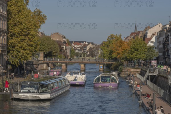 Tourist boat docks on the Ill and transports visitors who enjoy the view of Strasbourg's sights and trees in autumn. The scenery is peaceful and sunny. Bas Rhin, Alsace, France