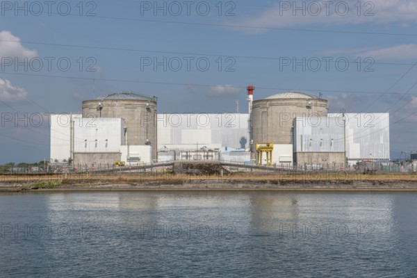 A large industrial nuclear power plant is located on a quiet water front. Two tall cylindrical structures dominate the skyline, with overhanging power lines and distant clouds that ennoble the blue sky. Fessenheim, Haut Rhin, Alsace, France