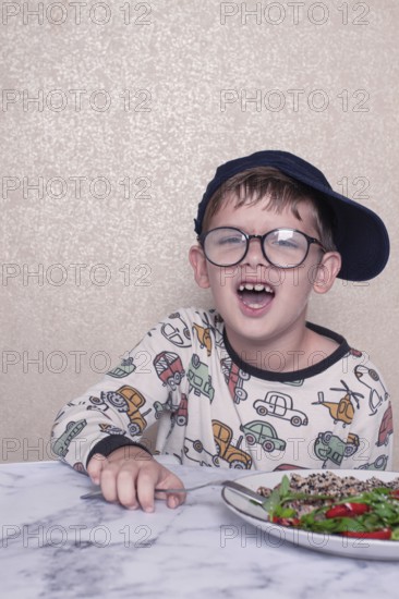 Child reacts dramatically to a salad served on a marble table at home during lunchtime, A little boy in glasses, and a cap, makes a funny face, sitting at a marble table. A salad is placed in front of him, demonstrating, his humorous reaction, during lunch