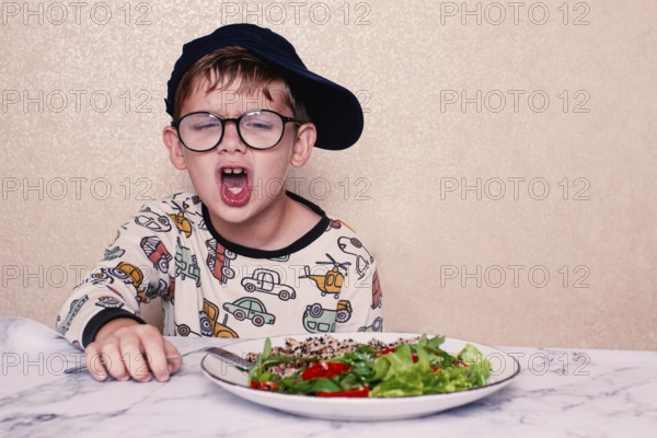 Child reacts dramatically to a salad served on a marble table at home during lunchtime, A little boy in glasses, and a cap, makes a funny face, sitting at a marble table. A salad is placed in front of him, demonstrating, his humorous reaction, during lunch