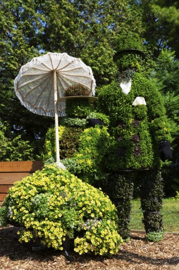 Live plant sculpture of Victorian man standing and woman with sun shade umbrella sitting on wooden park bench in summer, Ile des Moulins, Old Terrebonne, Quebec, Canada
