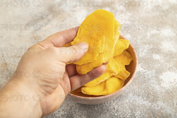 Dried Mango in ceramic bowl with hand on brown concrete background. Side view, close up. healthy food, minimalism