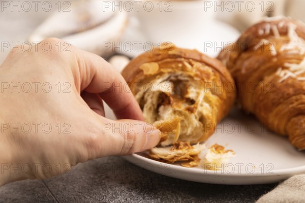 Croissant on white plate with hand on brown concrete background and linen textile, cup of coffee, side view, close up, selective focus