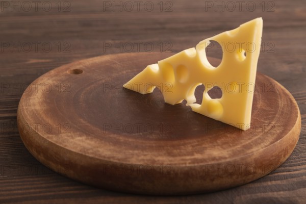 Piece of cheese with holes on cutting board on brown wooden background, side view, close up, minimalism