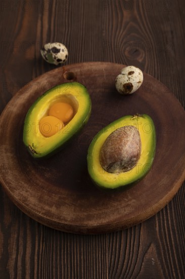 Two Avocado halves with pit and Egg yolk on round wooden cutting board on brown wooden, copy space, minimalism, low key, side view, close up