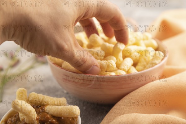 Corn flakes sticks with hand with caramel in ceramic bowl on gray concrete background and orange linen textile. Side view, close up