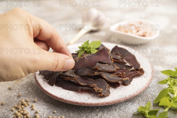 Armenian Basturma dried meat with hand on plate with pepper and herbs on brown concrete background. Side view, close up, selective focus