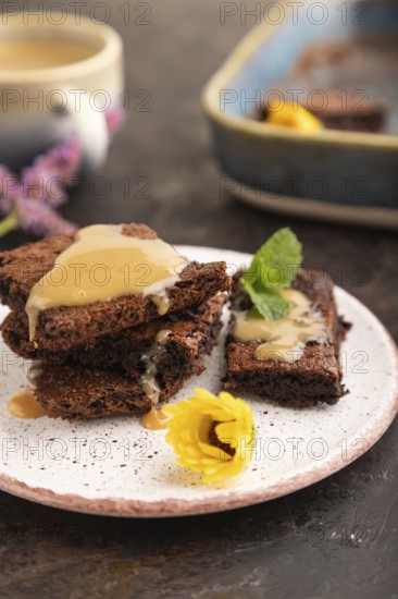 Chocolate brownie with caramel sauce with a cup of coffee on black concrete background. side view, close up, selective focus