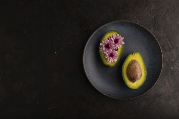 Two Avocado halves with pit and Chrysanthemums on blue plate on black concrete background, top view, flat lay, copy space, minimalism, low key