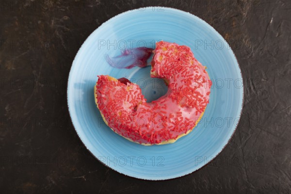Bitten Pink Donut with sprinkles on blue ceramic plate on black concrete background, top view, flat lay, copy space, minimalism