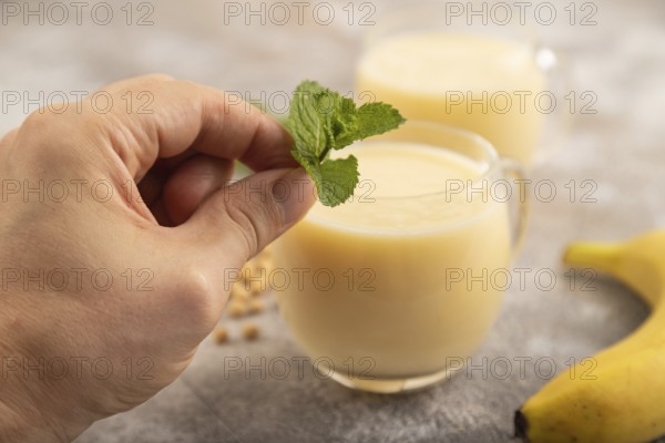 Organic non dairy banana and soy milk in glass with hand on brown concrete background. Vegan healthy food concept, side view, close up, selective focus