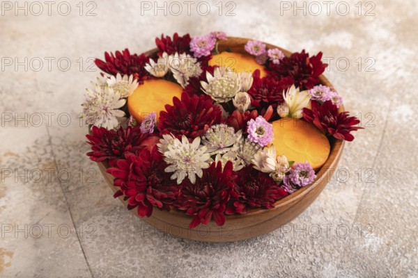 Wooden bowl with carrot slices and red Chrysanthemum flowers, Astrantia flowers, flower salad on brown concrete background, side view, close up, minimalism