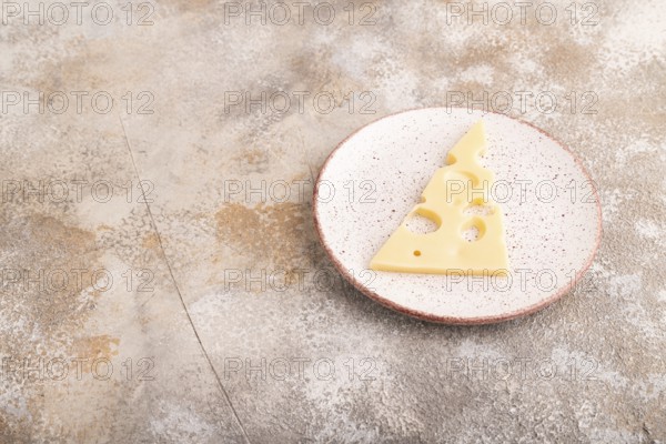 Piece of cheese with holes on ceramic plate on brown concrete background, side view, copy space, minimalism