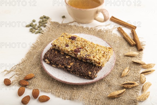 Granola bars with caramel, nuts, flakes in ceramic plate on white wooden background, beige linen napkin, cup of green tea. Side view, close up