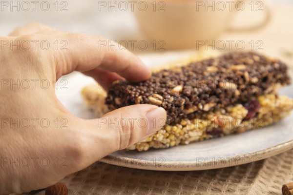 Granola bars with caramel, nuts, flakes in ceramic plate on brown concrete background with hand, beige linen napkin, cup of green tea. Side view, close up, selective focus