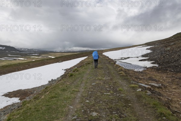 Hiker with rucksack and blue raincoat on a lonely plateau with snowfields, low clouds, Kongevegen or King's Road, Olav's Way or Olavsleden, Dovrefjell, Oppdal municipality, Trøndelag county, Norway