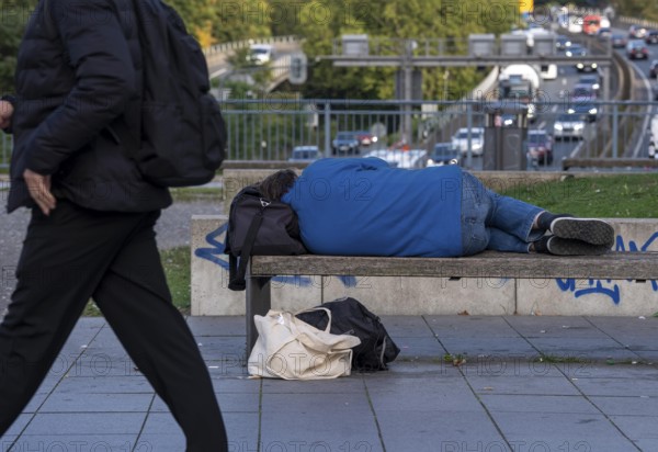 Man sleeping on a bench, travelling bag serves as pillow, in the city centre, passers-by walk by