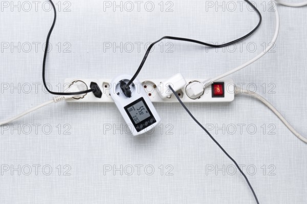Top view of a power strip with on/off switch and various inserted plugs, cables and an electricity cost meter on a white Resopal panel