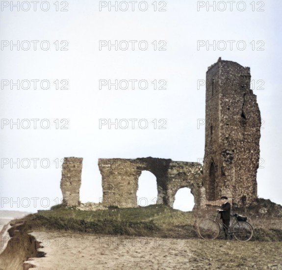 Colorised magic lantern slide of boy with bicycle standing by ruins of All Saints church, Dunwich, Suffolk, England, UK c 1900