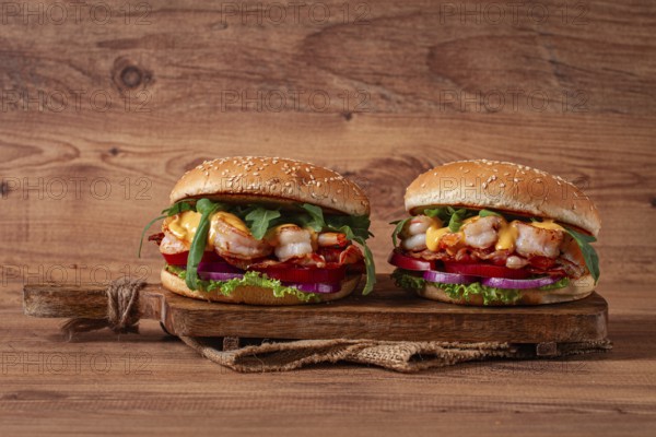 Two shrimp burgers and buns with sesame seeds, bacon, lettuce, tomatoes and onions, on a wooden board, rustic style