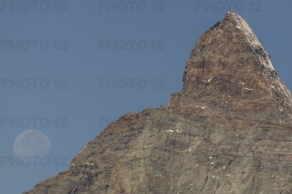 The Matterhorn mountain peak rises steeply against a bright blue sky. The full moon glistens gently above, creating a marvellous view in the serene evening hours. Matterhorn, Zermatt, Valais, Alps, Swiss