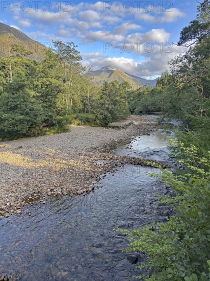 A tranquil river flows through a wooded landscape with mountains in the background and a clear blue sky, Glen Nevis, Highlands, Fort William, Scotland, United Kingdom