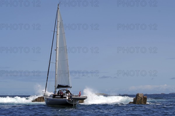 A sailing boat navigates through rocky sea with blue sky in the background, Isles of Scilly, Cornwall, United Kingdom