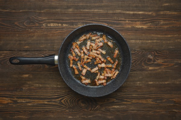 Sizzling strips of bacon fry in a black skillet, releasing savoury aroma and fat on rustic wooden tabletop