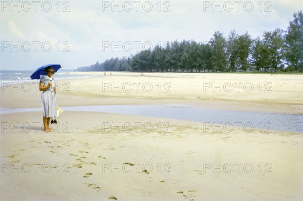 British woman holding umbrella standing on sandy beach south of estuary, Rompin, East coast, Pahang, Malaya, Malaysia, south east Asia 1964